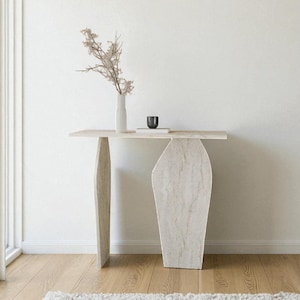Op de afbeelding: Een moderne, lichtgekleurde console met een rechthoekig blad en twee uniek gevormde poten. Een witte vaas met gedroogde bloemen en een kleine zwarte beker staan op de tafel. De tafel is gemaakt van steen of een vergelijkbaar materiaal.