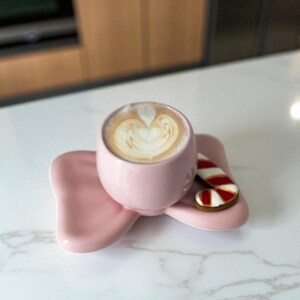 May include: A pink ceramic cup filled with a latte, resting on a matching saucer. A small gingerbread cookie shaped like a candy cane with red and white stripes sits on the saucer. The coffee has a heart-shaped design on top.