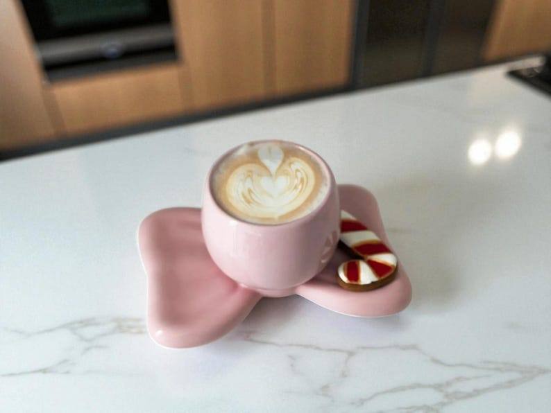 May include: A pink ceramic cup filled with a latte, resting on a matching saucer. A small gingerbread cookie shaped like a candy cane with red and white stripes sits on the saucer. The coffee has a heart-shaped design on top.