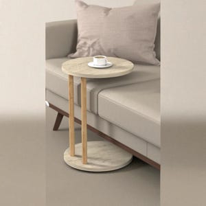 May include: A small, round side table with a light-coloured marble top and base, supported by three wooden legs. A white teacup and saucer sit on the table. The table is next to a beige sofa with a matching cushion.