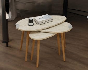 Puede incluir: Un juego de tres mesas nido con tapas ovaladas de color claro y patas de madera. Las mesas están dispuestas en una configuración apilada, con un pequeño objeto en blanco y negro y una pila de libros encima.