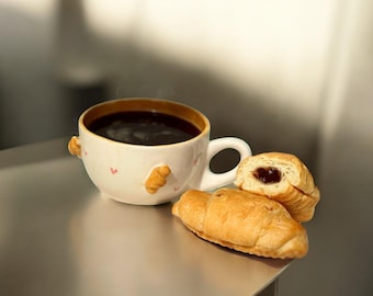 Puede incluir: Una taza de café de cerámica blanca con borde marrón y asa, llena de café oscuro. Dos pequeñas decoraciones en forma de croissant están adheridas a la taza. Dos cruasanes dorados, uno cortado para revelar un relleno oscuro, están junto a la taza.