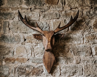 Può includere: Una scultura da parete a forma di testa di cervo intagliata in legno marrone scuro, caratterizzata da corna prominenti e una finitura liscia e lucida. L'oggetto presenta linee geometriche e una base appuntita che mette in risalto le venature.