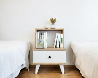May include: A modern nightstand with a white drawer and a light wood frame. The nightstand has a shelf with books, a small vase with dried flowers, and a small bowl. The nightstand is supported by white legs.