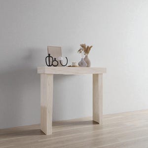 Könnte beinhalten: Eine hellfarbene Holzkonsolentisch mit rechteckiger Platte und vier quadratischen Beinen. Der Tisch ist mit einem Spiegel, zwei kleinen Vasen und einem dekorativen Kerzenhalter dekoriert. Der Hintergrund ist eine weiße Wand und ein Holzboden.