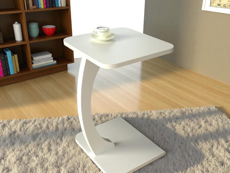 Pu&ograve; includere: Un tavolino a C bianco e moderno con un piano quadrato e un supporto curvo. Una tazzina e un piattino sono appoggiati sul tavolo.