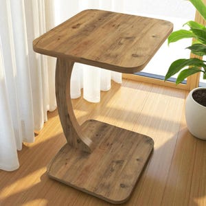 Pu&ograve; includere: Un tavolino in legno con piano e base quadrati, collegati da un supporto curvo. Il tavolo &egrave; realizzato in legno marrone chiaro e poggia su un pavimento in legno. Sullo sfondo &egrave; visibile una pianta in vaso.