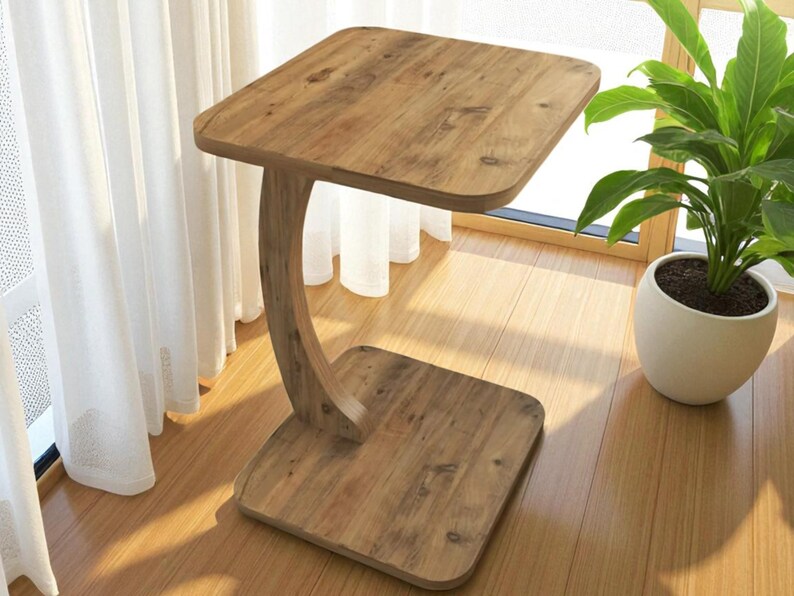 Pu&ograve; includere: Un tavolino in legno con piano e base quadrati, collegati da un supporto curvo. Il tavolo &egrave; realizzato in legno marrone chiaro e poggia su un pavimento in legno. Sullo sfondo &egrave; visibile una pianta in vaso.