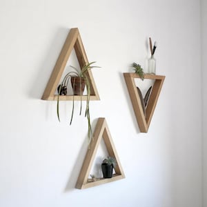 以下が含まれることがあります： 白い壁に三角形の棚が3つ取り付けられています。棚は、上の棚に小さな鉢植えの植物、下の棚に小さな鉢植えの植物がある以外は空です。