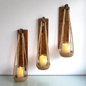 Puede incluir: Tres candelabros de pared. Cada uno presenta un globo de vidrio transparente que sostiene una vela encendida, suspendida por una cuerda tejida de una tabla de madera. Las tablas de madera están teñidas de marrón oscuro y montadas en una pared blanca.