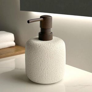 Könnte beinhalten: Ein weißer, strukturierter Seifenspender aus Keramik mit einer dunkelbraunen Pumpe und einem Ausguss. Der Spender hat eine abgerundete Form und steht auf einer weißen Oberfläche. Minimalistisches und modernes Design, für das Badezimmer.