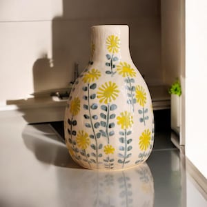 Könnte beinhalten: Eine weiße Keramikvase mit einem Blumenmuster. Die Vase hat einen runden Körper und einen schmalen Hals. Die Blumen sind gelb und grün.