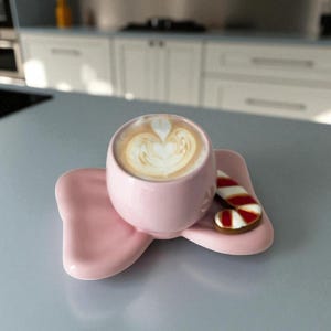 May include: A pink coffee cup with a heart-shaped design in the foam, resting on a matching pink saucer. A candy cane cookie sits beside the cup. The set is on a light gray surface.