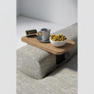 May include: A wooden side table with a black metal base attached to a couch. The table has a white ceramic cup, a small black clock, and a white bowl filled with peanuts.