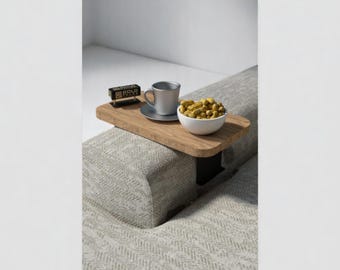 Op de afbeelding: Een houten bijzettafel met een zwarte metalen basis bevestigd aan een bank. De tafel heeft een witte keramische beker, een kleine zwarte klok en een witte kom gevuld met pinda's.