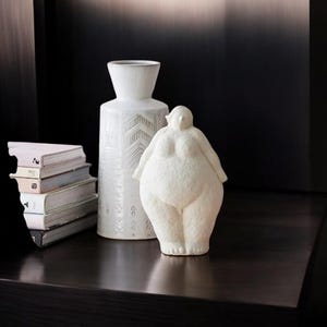 Puede incluir: Jarrón de cerámica blanca con un patrón geométrico y una escultura de cerámica blanca de una mujer sobre una superficie de madera oscura.