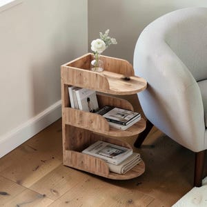Op de afbeelding: Een houten bijzettafel met drie gebogen planken, met boeken en een opgevouwen deken. Bovenop staat een vaas met witte bloemen. De tafel staat naast een lichtgrijze fauteuil. De vloer is van hardhout.