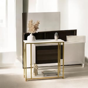 Puede incluir: Una mesa auxiliar moderna blanca y dorada con tapa de mármol. La mesa tiene una estructura de metal dorado y un estante inferior. Está decorada con un jarrón blanco con flores secas, una escultura de mano de cerámica y botellas decorativas. La mesa mide 76 cm de largo, 30 cm de ancho y 76 cm de alto.