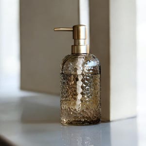 May include: A decorative amber glass soap dispenser with a textured surface and a gold-colored pump. The dispenser is cylindrical with a rounded top and is placed on a white surface.
