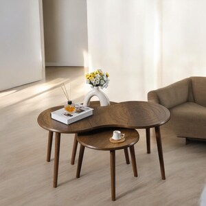May include: A pair of wooden nesting coffee tables. The larger table has a distinctive, curved design, and the smaller table is round. A white vase with flowers, a book, and a cup are on the tables. The tables have dark brown legs.