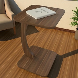 May include: A dark brown, C-shaped side table with a square top and base, and a curved support. A white book with text is on the table. The table is made of wood and is next to a beige armchair and a potted plant.
