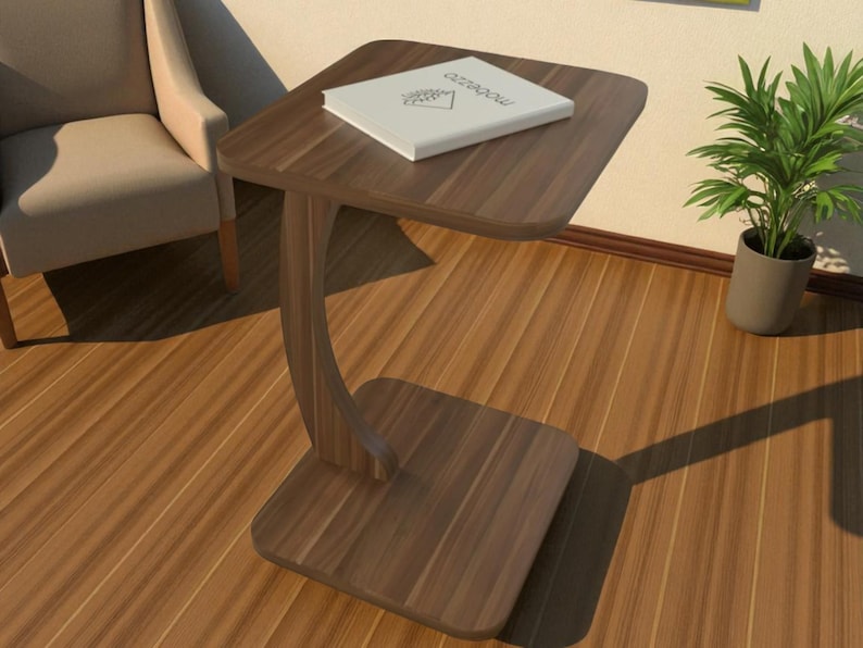 Pu&ograve; includere: Un tavolino laterale a forma di C, marrone scuro, con piano e base quadrati e supporto curvo. Un libro bianco con testo &egrave; sul tavolo. Il tavolo &egrave; in legno ed &egrave; accanto a una poltrona beige e una pianta in vaso.