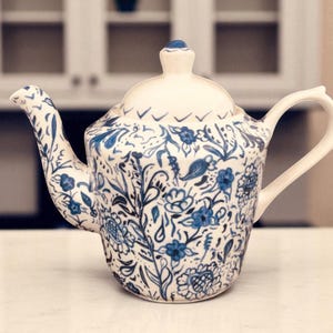 May include: White ceramic teapot with a lid and handle, adorned with a blue floral pattern. The teapot features a spout and a small knob on the lid. The design includes flowers, leaves, and vines, creating a classic aesthetic.