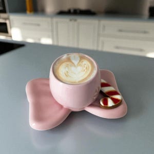 May include: A pink ceramic coffee cup with a heart-shaped latte art design, resting on a matching bow-shaped saucer. A candy cane cookie sits beside the cup. The items are on a gray surface.