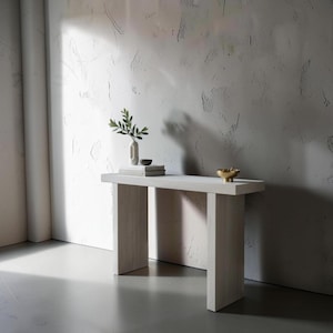 May include: A light-colored wooden console table with a rectangular top and two thick, rectangular legs. The table is decorated with a small vase with greenery, a small bowl, and a stack of books. A small gold-colored bowl sits on the table.