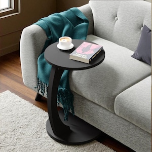May include: A black, round side table with a curved support, holding a book and a cup of coffee. A teal throw blanket with fringe is draped over a gray sofa. The table is on a white rug.