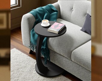 Puede incluir: Una mesa auxiliar redonda negra con soporte curvo, con un libro y una taza de café. Una manta turquesa está sobre un sofá gris. La mesa está sobre una alfombra blanca. La mesa mide unos 60 cm de altura.