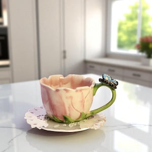 Könnte beinhalten: Eine rosa-weiße Keramik-Teetasse in Form einer Blume, mit einem grünen Henkel und einem blauen Schmetterlingsakzent. Die Teetasse steht auf einer weißen, spitzenbesetzten Untertasse. Die Teetasse steht auf einer weißen Arbeitsplatte.