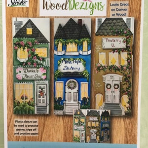 May include: A sheet featuring painted cottage boards with the text "Welcome Cottage Boards." The boards are in the shape of houses, with details like windows, doors, and floral accents. The text "WoodDezigns" is also visible.