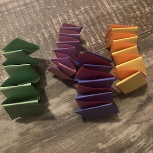 Puede incluir: Tres montones de papel doblado en tonos verdes, morados y naranjas. El papel está doblado en un patrón en zigzag, creando un efecto en capas. La disposición está sobre una superficie de madera.