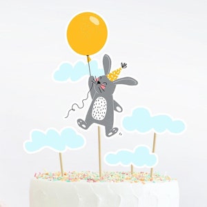 Könnte beinhalten: Ein weißer Kuchen mit einem grauen Hasen, der einen gelben Ballon hält und einen Partyhut trägt. Der Hase ist von blauen Wolken umgeben. Der Kuchen ist mit bunten Streuseln verziert.