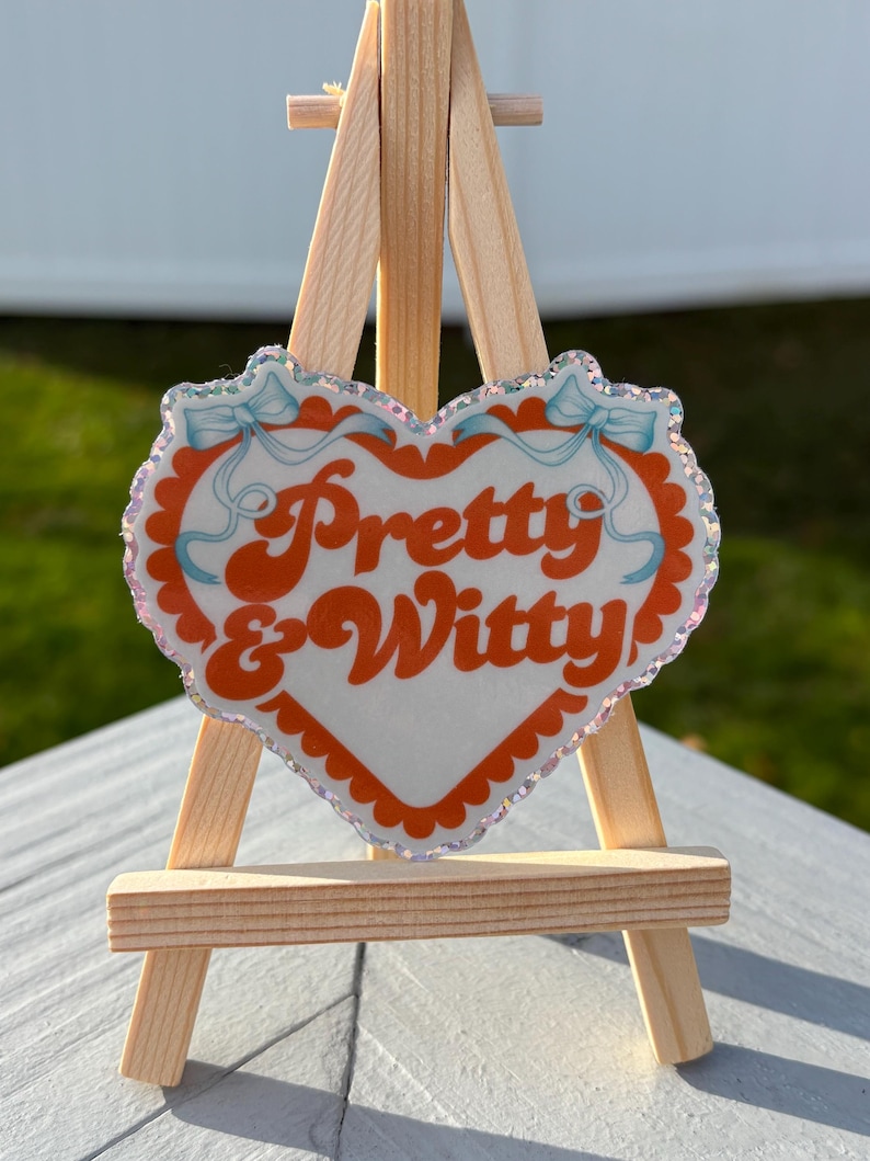 May include: A heart-shaped sign with the words "Pretty & Witty" in orange script. The sign has a white background with an orange border and light blue bows. It is displayed on a small wooden easel.