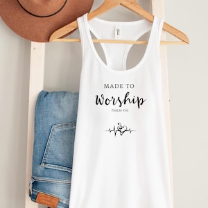 K&ouml;nnte beinhalten: Wei&szlig;es Tanktop mit dem Text "MADE TO Worship" und Psalm 95:6 in Schwarz gedruckt. Eine Grafik mit Noten und Herzschlag befindet sich unter dem Text. Ein brauner Hut und blaue Jeans sind im Hintergrund.
