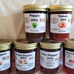 May include: Six glass jars of homemade pepper jelly and jam. The jars are labeled with the flavors: Onion Pepper Jelly, Peach Pepper Jelly, Hot Pepper Jelly, Strawberry Pepper Jelly, Pepper Jelly, and Tomato Jam. Each jar has a gold lid and a white label with a red and black logo.