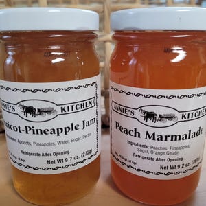 May include: Two glass jars of homemade jam, one with apricot-pineapple jam and the other with peach marmalade. Both jars have white lids and labels with the text "Annie's Kitchen" and the ingredients listed.