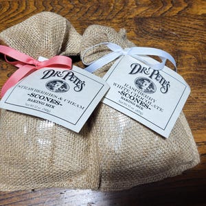 May include: Two burlap bags of Dr. Pete's scone mix. One bag is labeled "Strawberries & Cream" and tied with a pink ribbon. The other bag is labeled "Raspberry White Chocolate" and tied with a white ribbon. Each bag has a printed label.