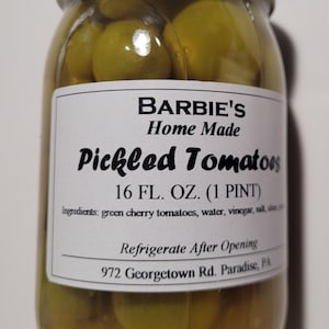 May include: A glass jar filled with green pickled cherry tomatoes. The label reads "BARBIE'S Home Made Pickled Tomatoes 16 FL. OZ. (1 PINT)". The jar is sealed with a white lid and the ingredients are listed on the label.