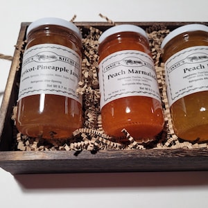 May include: A wooden gift box filled with brown shredded paper and three glass jars of jam. The jars are labelled "Apricot-Pineapple Jam", "Peach Marmalade", and "Peach Jam".