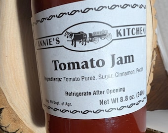 May include: A glass jar of Annie's Kitchen Tomato Jam with a white label featuring a horse-drawn wagon illustration. The label lists ingredients as tomato puree, sugar, cinnamon, and pectin, with a net weight of 8.8 oz. It states to refrigerate after opening.