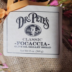 May include: A package of Dr. Pete's Classic Focaccia olive oil skillet bread. The label features the brand name and a graphic. The net weight is 13 oz (369 g). The package is wrapped in burlap and tied with an orange ribbon.