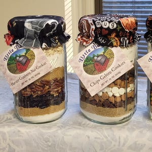 May include: Four glass jars filled with cookie ingredients, each with a fabric lid. The jars are labeled "Cranberry Chip Cookies", "Harvest Cranberry Nut Cookies", "Chips Galore Cookies", and "Peanut Butter Chocolate Jumbos".