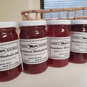 May include: Four glass jars of strawberry rhubarb jam with white lids and labels. The labels are white with black text and a black and white illustration of a horse-drawn wagon. The labels read "Annie's Kitchen" and "Strawberry Rhubarb Jam". The jars are arranged in a row on a white surface.