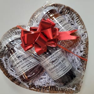 Peut inclure: Un panier en osier en forme de cœur rempli de trois pots en verre de confiture. Les pots sont enveloppés de cellophane et attachés avec un ruban rouge. Les étiquettes sur les pots indiquent "Strawberry Rhubarb", "Elderberry Jelly" et "Marmalade".