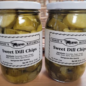 May include: Two glass jars of Annie's Kitchen Sweet Dill Chips. Each jar is filled with sliced pickles and topped with a white lid. The labels read "Sweet Dill Chips" and list ingredients. Each jar contains 16 oz.