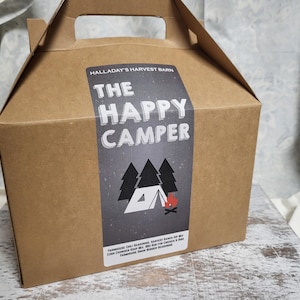 May include: A brown cardboard box with a handle, featuring a label that reads "THE HAPPY CAMPER." The label includes illustrations of pine trees, a tent, and a campfire. The box is designed for carrying items.