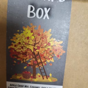 May include: A rectangular label with the text "The Orchard Box" above an illustration of an apple tree with a ladder. The label lists the following mixes: Apple Crisp, Caramel Apple Dip, Cider Mulling, Pumpkin Cheesecake, and Maple Cheesecake.